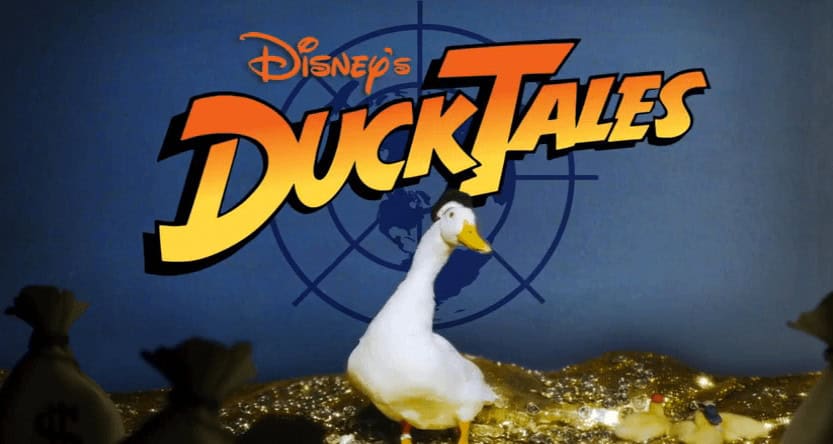 A real duck poses atop a pile of gold coins and money bags beneath the DuckTales logo on a vibrant blue backdrop, evoking the whimsical adventure and nostalgia that make Disney parks and DuckTales attractions so captivating.