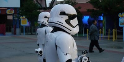 Two people dressed as Star Wars stormtroopers walk outside at Disney's Hollywood Studios, possibly near the Launch Bay, with another person in the background. Trees and colorful signs add to the Star Wars Experience at this Disney World park, Disney's Hollywood Studios. Disney World Closes Star Wars Location; Removes Wait Time Sign and More