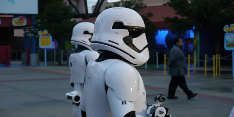 Two people dressed as Star Wars stormtroopers walk outside at Disney's Hollywood Studios, possibly near the Launch Bay, with another person in the background. Trees and colorful signs add to the Star Wars Experience at this Disney World park, Disney's Hollywood Studios. Disney World Closes Star Wars Location; Removes Wait Time Sign and More