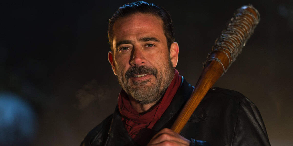 A person with a beard, reminiscent of Negan from *The Walking Dead*, holds a spiked baseball bat over their shoulder. Dressed in a leather jacket and red scarf, they stand in a dimly lit setting that feels like it's straight out of a TV spot.
