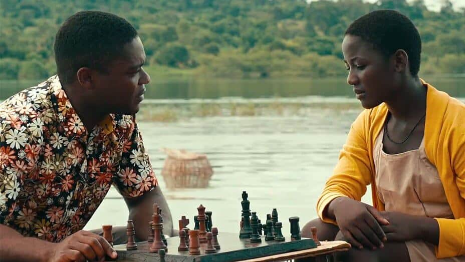 Two people sit across from each other playing a live chess game on a small table. They are outdoors near a body of water with lush greenery in the background. The person on the left wears a floral shirt, and the person on the right, channeling Queen of Katwe vibes, sports a yellow cardigan.