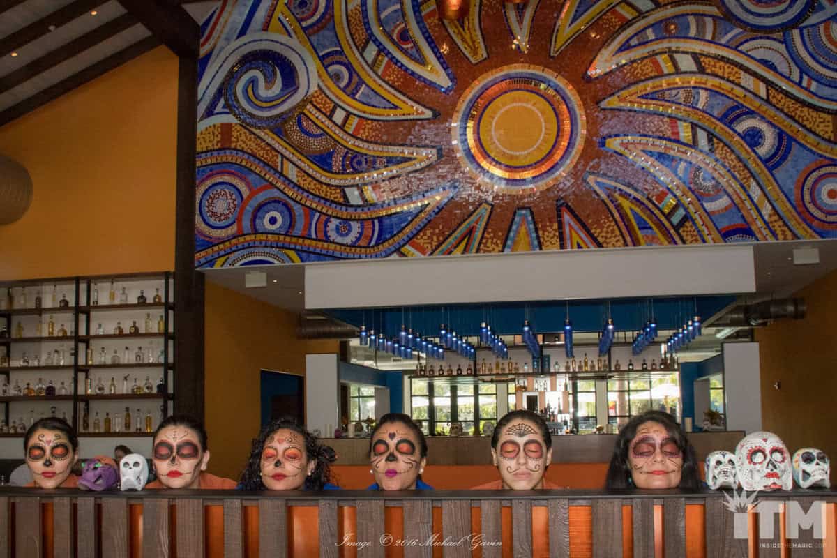 Guests with vibrant Dia De Los Muertos skull makeup enjoy the festive atmosphere at Frontera Cocina in Disney Springs, seated beneath a lively mosaic mural and shelves of colorful bottles, embodying the spirited celebration in this popular Disney dining spot.