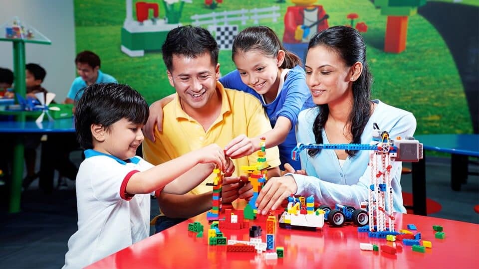 A family of four is enjoying a playful and educational activity by building with colorful LEGO blocks at a table. The father and daughter are smiling and working on a structure, while the mother and son engage with other pieces, reminiscing about their first visit to the LEGOLAND hotel in the Middle East.