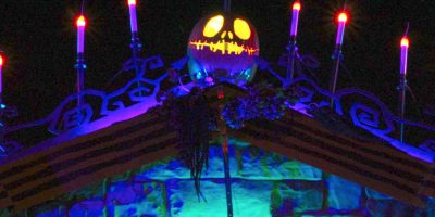 A glowing Jack-o'-lantern with a sinister face sits atop a rooftop, surrounded by lit candles in the dark, creating a spooky, Haunted Mansion Holiday scene perfect for any Disney Fan's at-home imagineering.