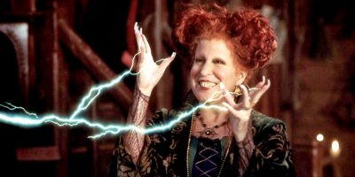 A woman with curly red hair and a green dress magically conjures blue lightning from her hands, smiling mischievously in a dimly lit, rustic room—reminiscent of Bette Midler’s iconic witch in a Disney Channel Hocus Pocus remake.