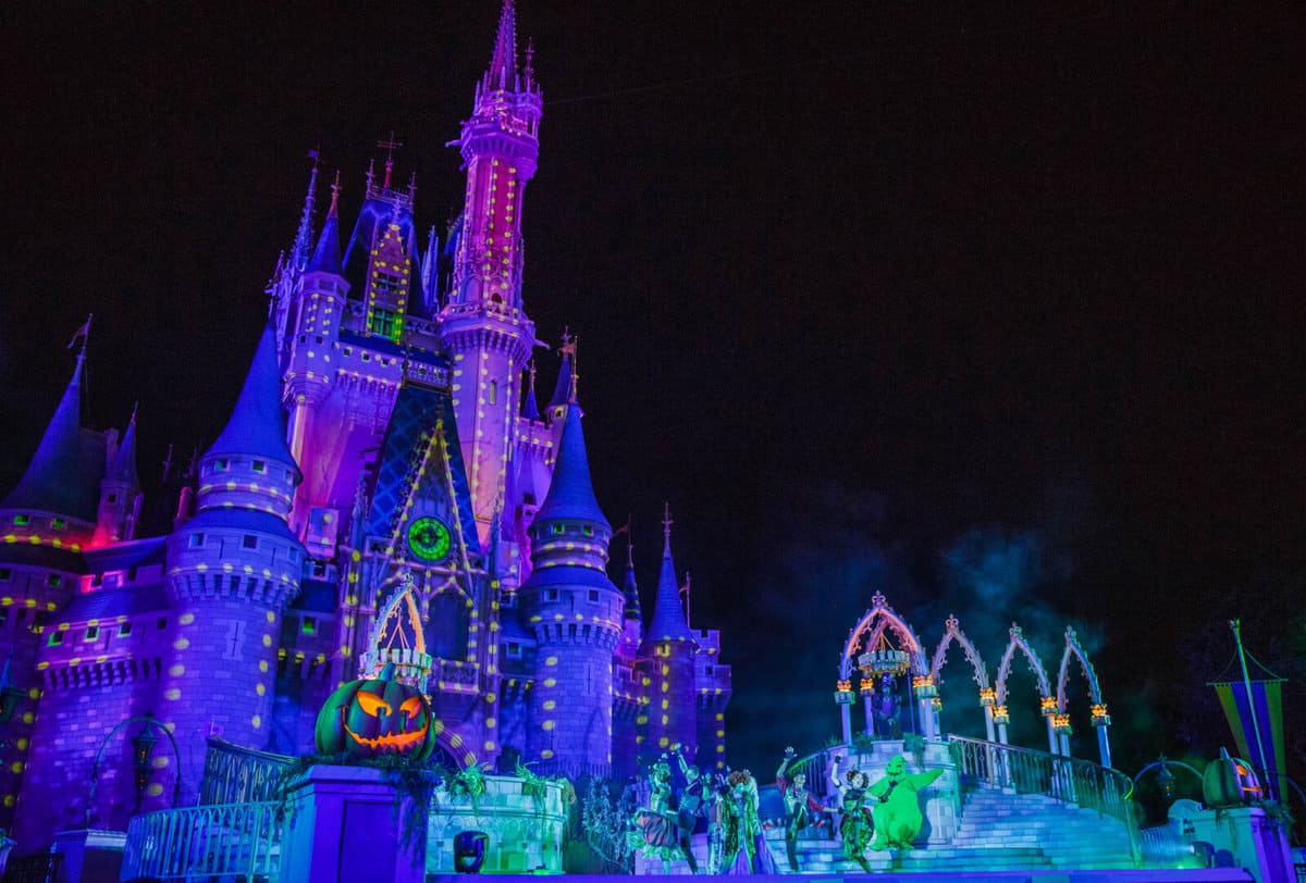 A brightly lit castle at night is decorated with colorful lights and Halloween-themed projections, including a large glowing jack-o'-lantern, while Hocus Focus performers gather near the castle’s entrance at a Disney park.