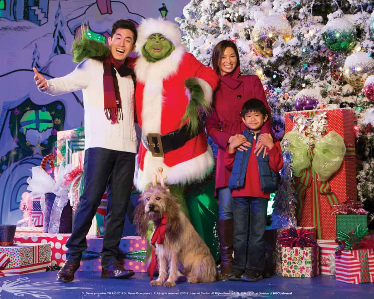 The Grinch poses with a joyful family and their dog beside a festive Christmas tree and stacks of colorful presents, bringing holiday cheer to Universal Studios Hollywood guests.