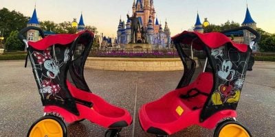 Two empty red and black strollers, featuring images of Mickey and Minnie Mouse, are parked facing each other on an empty path in the Magic Kingdom. The strollers are positioned in front of a castle with blue rooftops and a statue, set against a clear sky in the amusement park.