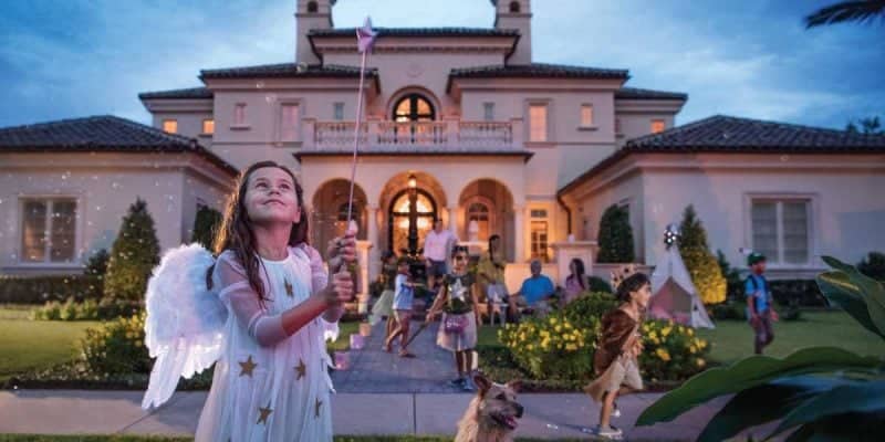 A young girl dressed as an angel with wings and a wand stands in front of a large, elegant house resembling something from Disney World, with children in various costumes playing around her. The scene is festive and magical, framed by a golden oak tree and a dog in the foreground as the sky turns to dusk.