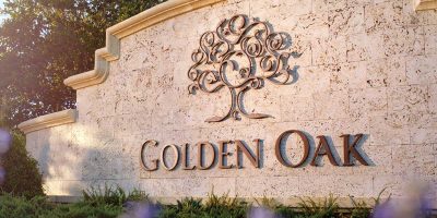 A stone wall with a decorative tree emblem and the text "GOLDEN OAK" below it, set against a green foliage background, evokes the charm of Disney World. The wall has a beige texture, and the letters and tree symbol are in a dark bronze color.