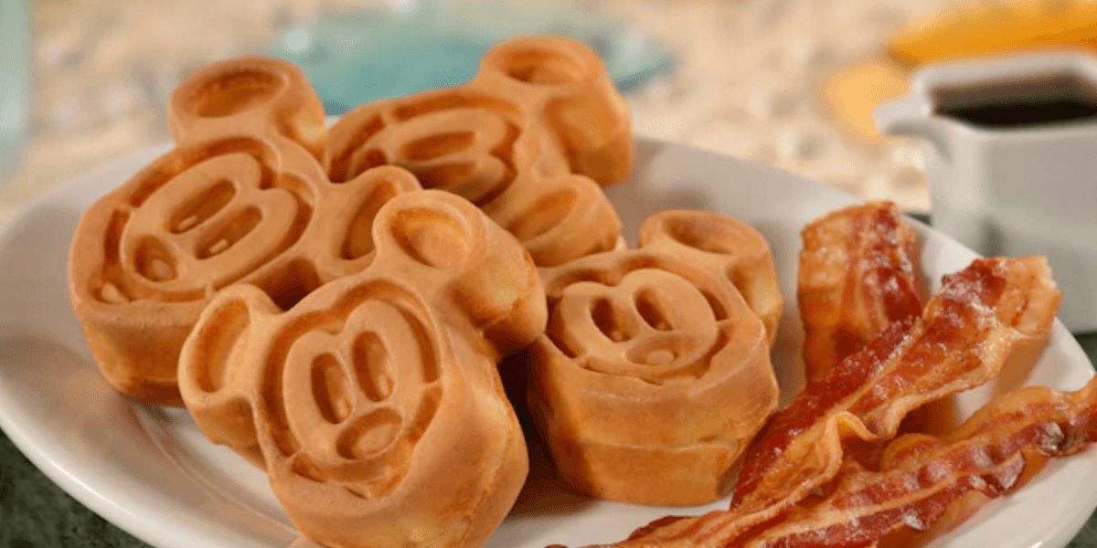 A white plate holds three waffles shaped like cartoon mouse faces, a fun treat often enjoyed with the Disney Dining Plan, accompanied by several strips of crispy bacon. A syrup container is partially visible in the background at Disneyland California, a Disney park.