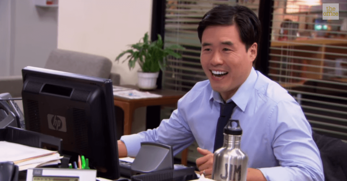 Randall Park sitting at a table with a laptop and smiling at the camera