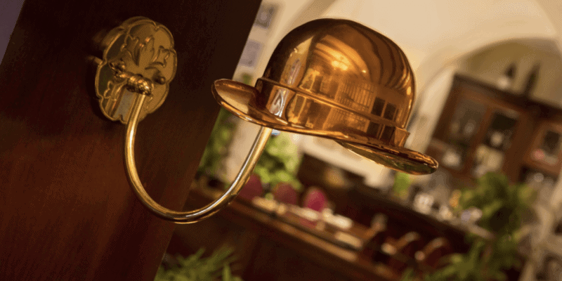 A wall-mounted lamp designed to look like a metallic bowler hat casts a warm golden glow, reminiscent of the iconic Hollywood Brown Derby in Disney world. The lamp curves outward from the wall against a background featuring blurred furniture and greenery, suggesting a cozy indoor setting.