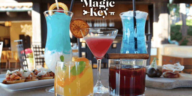 Five colorful cocktails in different glasses are displayed on an outdoor table at Magic Key Terrace Disneyland, with appetizers in the background. The text "Magic Key" appears above the drinks in a stylized font.