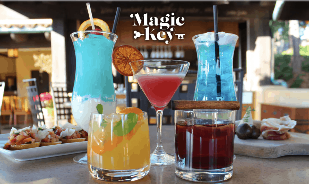 Five colorful cocktails in different glasses are displayed on an outdoor table at Magic Key Terrace Disneyland, with appetizers in the background. The text "Magic Key" appears above the drinks in a stylized font.