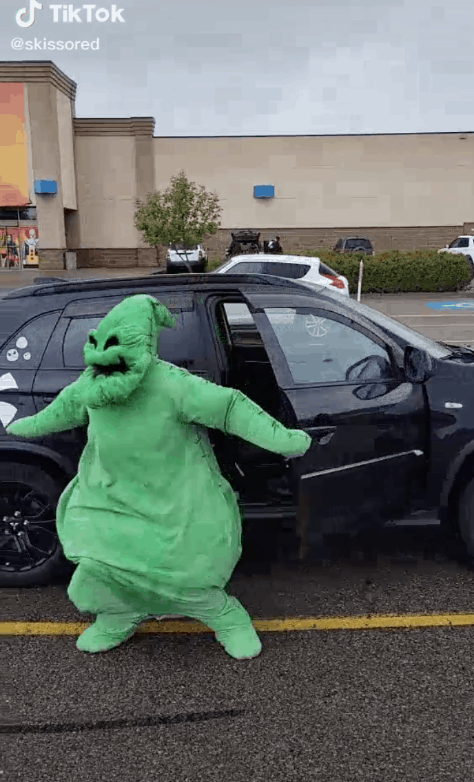 A Disney Villain electrifies the parking lot outside a sleek black SUV, swirling with wicked energy in a vivid green costume beneath cloudy skies, captivating fans with their dramatic performance.