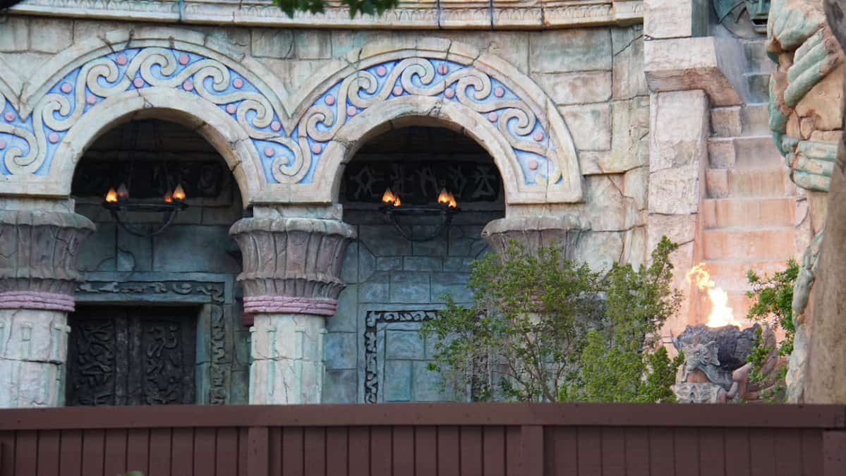 Iconic Universal Studios stone arches adorned with swirling blue designs and blazing torches flank a central flame, set before a rustic brown fence and lush green shrubbery, building excitement for the eagerly awaited reopening of this legendary attraction.