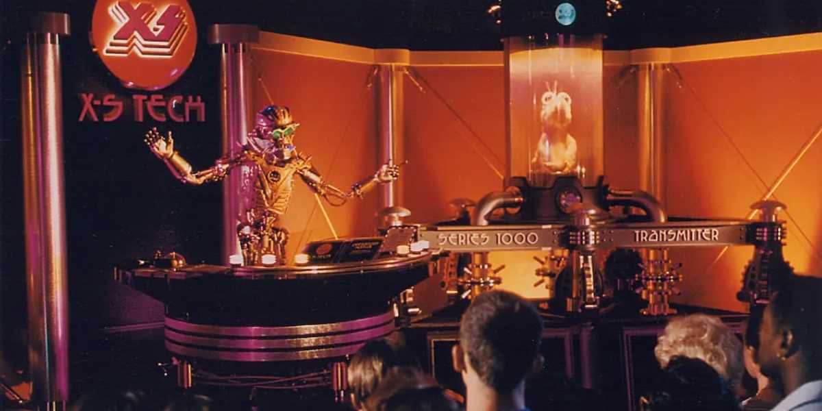 A futuristic scene unfolds with a robotic figure poised, arms outstretched, at a control panel labeled "Series 1000 Transmitter" during the mesmerizing X-S Tech exhibit. In the background, a cylindrical chamber ominously holds a creature inside, captivating onlookers like a thrilling Disney ride.