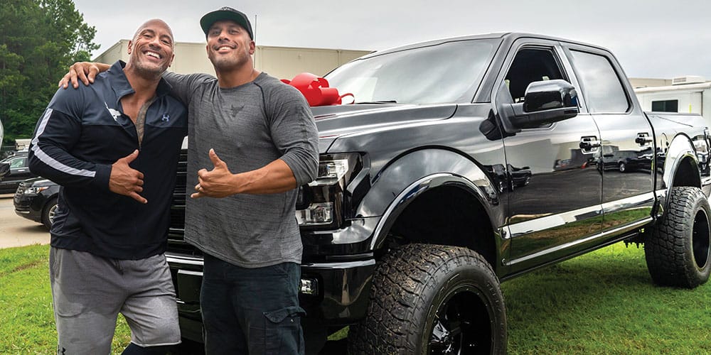 Two stunt performers beaming in front of a gleaming black pickup truck topped with a giant red bow, throwing up thumbs up as they celebrate their Stuntperson of the Year victory—looking like they’ve just wowed the crowd at Universal Studios with an action sequence straight out of a blockbuster.
