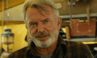 Sam Neill from the Dominion cast, rocking a gray beard and plaid shirt, stands inside an adventure-themed attraction with camping gear props behind him, channeling his signature rugged explorer vibe.