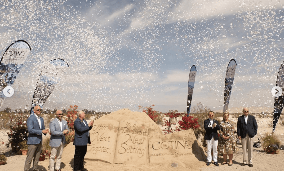 Six guests gather beside a sand sculpture announcing the grand opening of a new Disney Storyliving community, surrounded by fluttering white confetti and waving flags that evoke the magic and celebration found at Disney parks.