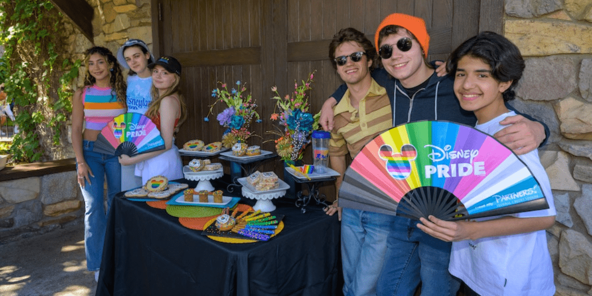 The Cast of 'Love Victor' displays Disney Pride fans in front of a table full of Pride snacks.