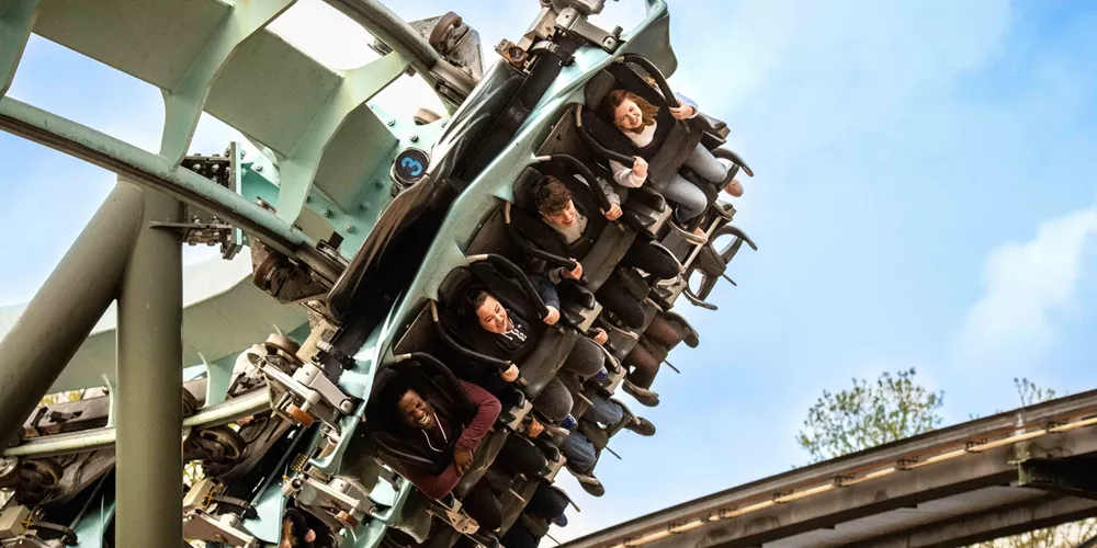 Guests, including a young girl with a prosthetic leg, experience the thrills of an inverted coaster at a theme park, their faces lit up with excitement and fear as they soar against the bright blue sky.