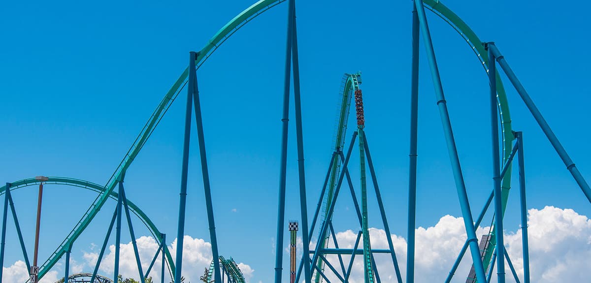 The park’s towering roller coaster, featuring striking teal and green tracks, twists and soars beneath a vivid blue sky dotted with fluffy clouds as crowds enjoy the excitement below.