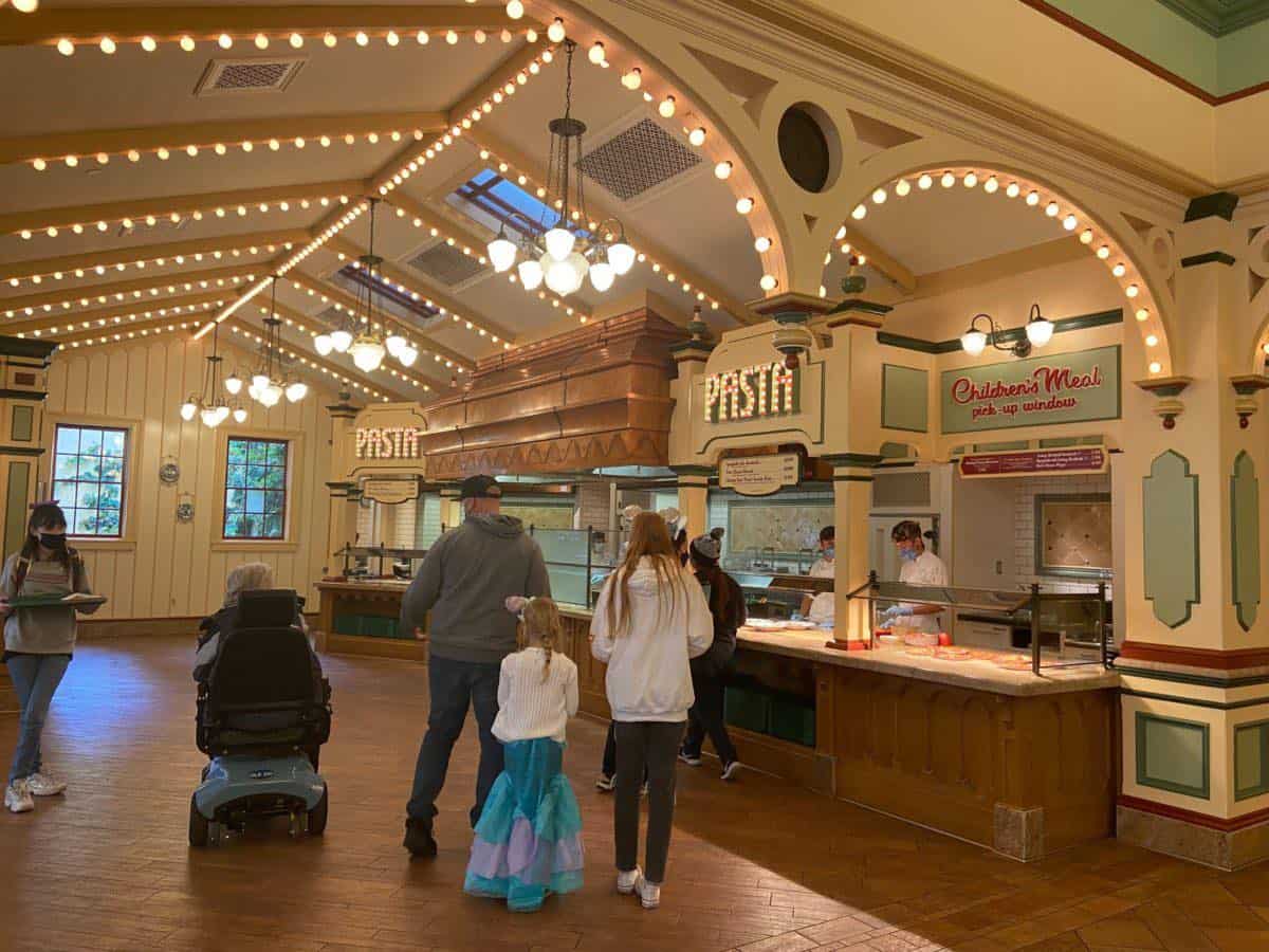 Guests explore a brightly lit cafeteria inside Disney California Adventure, passing a pasta station and a sign for kids’ meals on the right, experiencing one of the park’s hidden gem dining spots that offers a relaxing escape from typical Disneyland Resort crowds.