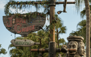 A wooden “Jingle Cruise” sign, inspired by the Jungle Cruise New Version, hangs among palm trees, adorned with festive lights and greenery. Below is a green “Exotic” arrow sign and to the right, a carved stone face statue.
