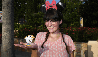 A girl with glasses, brown braids, and black Minnie Mouse ears looks at her hand, where Chip (a teacup) has been added in post-production.
