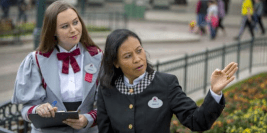 Two Cast Members in gray and black suits pointing in front of Sleeping Beauty Castle at Disneyland Park (Paris).