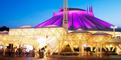 Nighttime view of the Space Mountain attraction at Disneyland, illuminated in purple and white lights. The structure features a futuristic design with a prominent pointed rooftop. Guests can be seen queuing under the brightly lit entrance canopy as Disney prepares to close for the day.