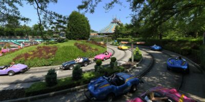 A colorful scene at Disneyland Paris, showing people driving miniature cars along a curvy track surrounded by lush greenery and neatly manicured bushes under a bright sky.