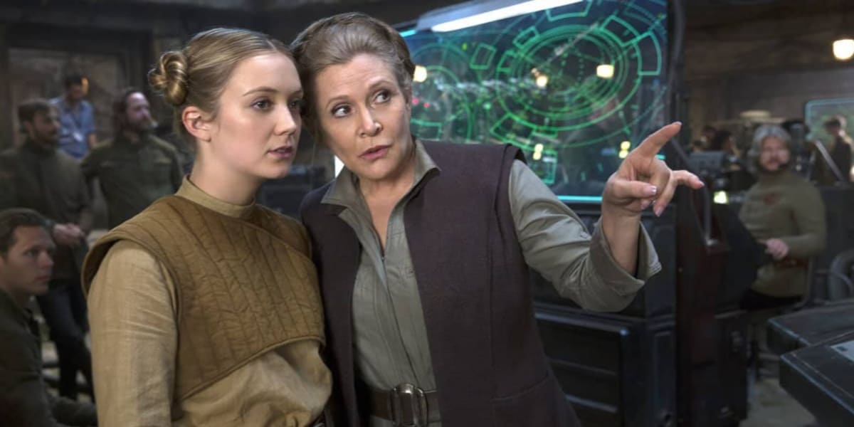 Two women in sci-fi attire are standing in a high-tech control room. The older woman, wearing a dark vest over a gray top, points at something on a digital screen, explaining it to the younger woman, who is dressed in a brownish outfit reminiscent of Carrie Fisher's iconic look. People work in the background.