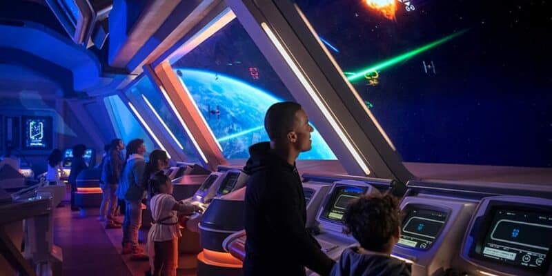 Guests at the new Disney hotel look out of a large window at a space battle simulation, featuring spaceships and neon blasts, with interactive consoles in the foreground.