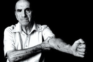 A black and white image of an elderly man showing his numerical inner arm tattoo. He survived a concentration camp.