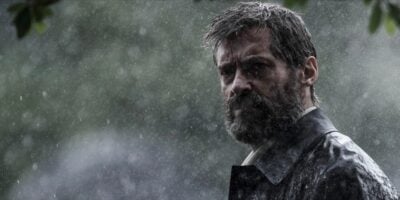 Hugh Jackman with a solemn expression stands in the rain, his wet hair and clothes sticking to him, with blurred green foliage of the X-Men training ground in the foreground.