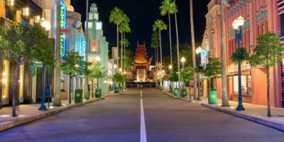 An empty street at night in Disney Hollywood Studios with illuminated art deco-style buildings and a replica of a famous theater facade in the distance.