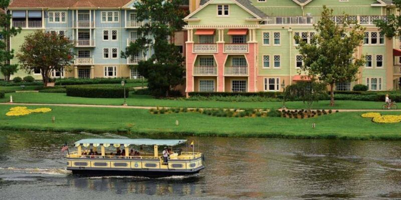 A charming riverboat filled with passengers cruises along a river in front of a row of colorful, multi-story buildings in a quaint residential area with lush green lawns, shortly before Disney suspends water