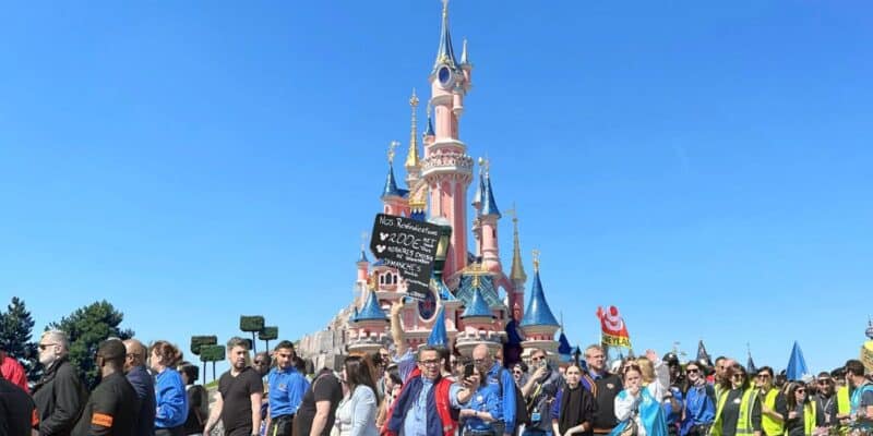 A group of people, some holding protest signs in French, are gathered in front of a colorful castle under a clear blue sky. The castle's pink spires and golden accents resemble a fairy-tale design. It appears to be the site of ongoing cast member strikes in Paris.