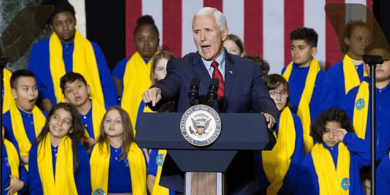Mike Pence speaks at a podium in front of about a dozen children in blue and yellow robes. 
