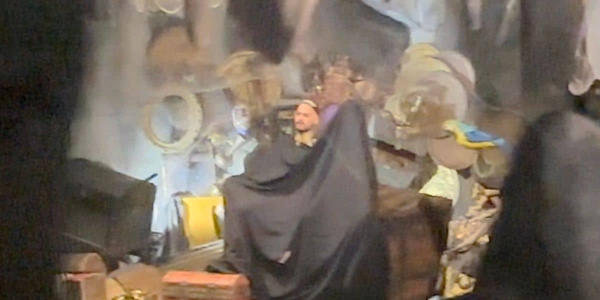 A mysterious figure, channeling the iconic pirate flair of Captain Jack Sparrow, sits in a dimly lit setting reminiscent of an attraction at a themed entertainment venue, draped in dark cloth and surrounded by an eclectic array of hats and props stacked on shelves and crates.