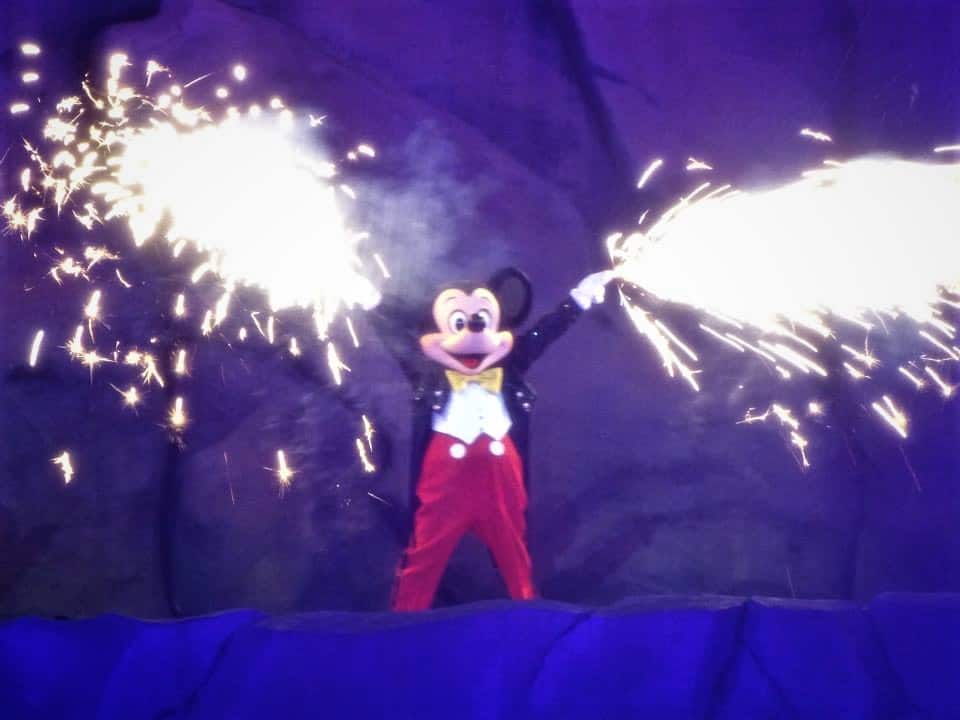 Mickey Mouse stands on stage with arms raised, shooting sparks from his hands against a dark, craggy backdrop, resembling a Fantasmic performance at night inside Disneyland Park.