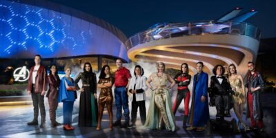 Group of diverse people dressed in futuristic costumes standing in front of the Avengers Campus building with illuminated blue lights and a sleek tram above.