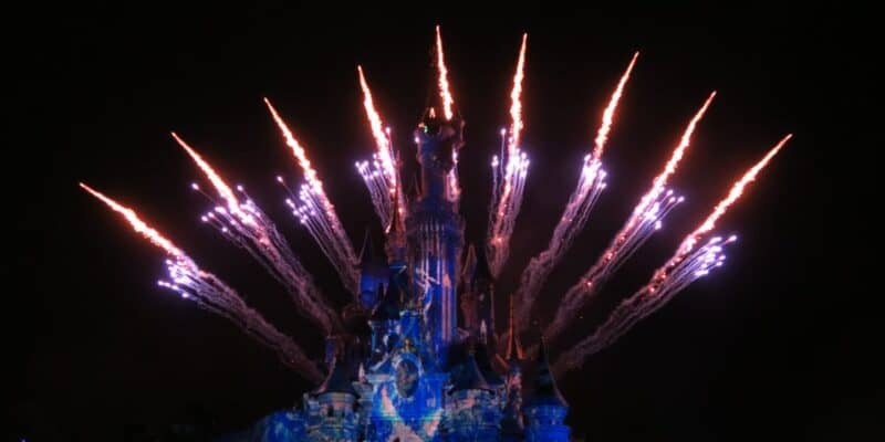 A colorful fireworks display illuminates the night sky behind a brightly lit, fairy tale castle with detailed blue and purple projections on its surface. The fireworks create a fan-like pattern, adding a magical atmosphere to the scene, enchanting Disneyland Paris guests.