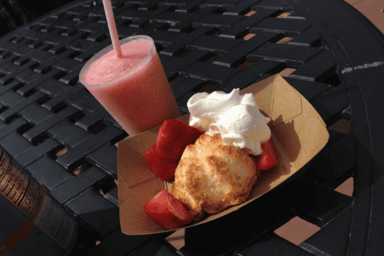 A desert pastry in a brown take-out plate, accompanied by strawberries and whipped cream. Alongside the desert is a pink slushy or smoothie textured beverage.