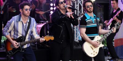 Three musicians perform on stage. The one in the middle, reminiscent of Nick Jonas, sings into a microphone while the others play guitars. They wear sunglasses and casual outfits. A drummer plays in the background as audience members cheer, unaware of any recent injuries affecting their perfection.