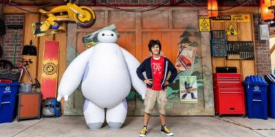 A young person stands in front of a workshop-themed backdrop with a large, white, inflatable robot character. The person is smiling with hands on hips. The workshop has red toolboxes, posters, and various equipment, creating a vibrant, industrial atmosphere that would captivate any Disney guest.