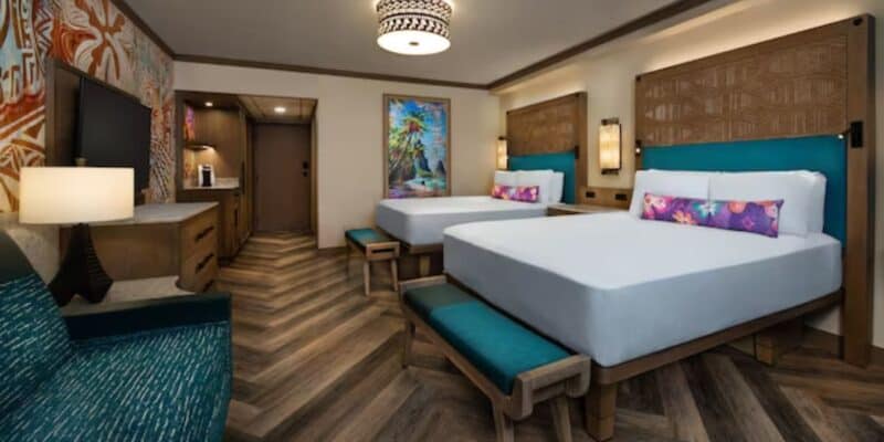 A hotel room with two beds featuring white linens and colorful pillows, a wall-mounted TV, a patterned carpet, a desk with a lamp, and tropical-themed decor. The room includes a vibrant painting, wooden furnishings, and soft lighting that creates an atmosphere where relaxation is rampant.